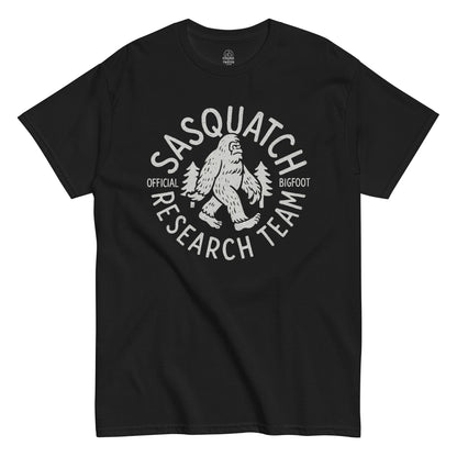 Sasquatch Research Team T-shirt featuring white Bigfoot walking between pine trees with bold text, on black background.