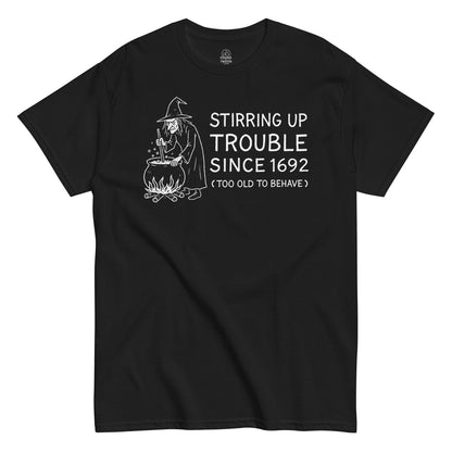 Funny witchcraft T-shirt with white artwork of a witch stirring a cauldron and text “Stirring Up Trouble Since 1692” on black fabric.