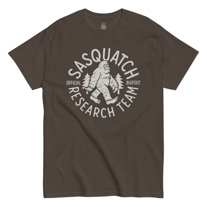 Sasquatch Research Team T-shirt showing white Bigfoot and pine trees design on dark brown background.