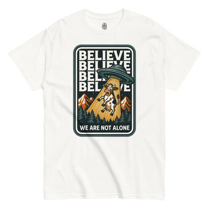 Funny UFO T-shirt with cartoon cow being abducted by a UFO, text “Believe – We Are Not Alone,” printed on white fabric
