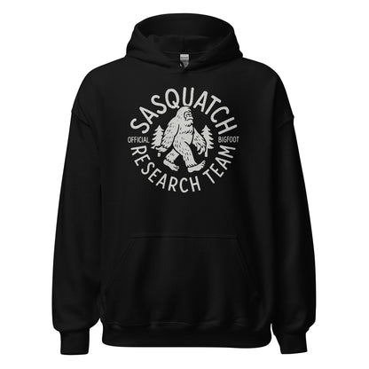 Black hoodie featuring the Sasquatch Research Team cryptid emblem in light grey, centered on chest, Bigfoot silhouette with trees, circular text layout, By Strange & Twisted